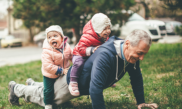 A photo of a playful grandfather and granddaughter. They are casually dressed and playing in the park. They exercise together. A grandfather is exercising while granddaughters are sitting on his back.