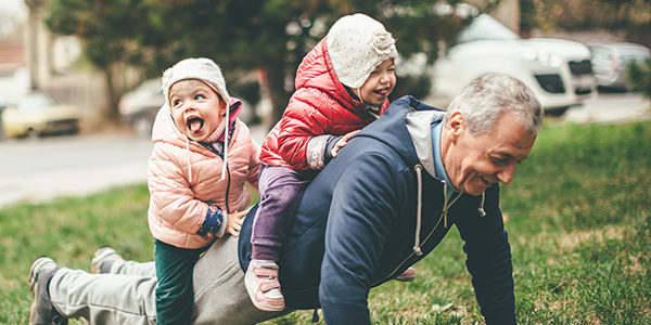 A photo of a playful grandfather and granddaughter. They are casually dressed and playing in the park. They exercise together. A grandfather is exercising while granddaughters are sitting on his back.